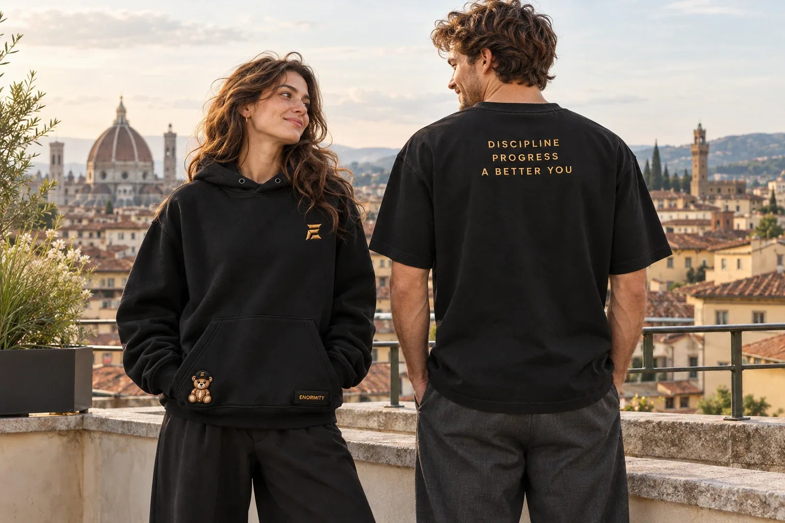 Žena v černé mikině Teddy Hoodie se zlatým E, medvídkem a štítkem ENORMITY, vedle muž zezadu v černém tričku s nápisem Discipline, Progress, A better you; terasa nad Florencií (vizualizace)